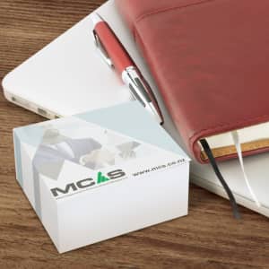 A white memo cube note pad with 400 leaves, positioned on a wooden desk alongside a red pen and notebook.