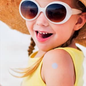 A child in sunglasses and a sun hat smiles, with a small sticker on their arm, wearing a yellow top.