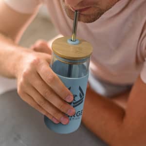 A light blue glass tumbler with a bamboo lid and stainless steel straw, held by a person. It features a logo.