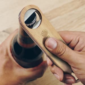 A drink bottle opener made of wood and metal, being used to open a dark beverage bottle. It features a logo.