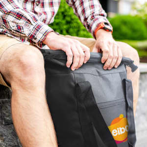 A black roll top cooler bag held by a person sitting down, featuring a logo on the front.