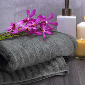 A grey bath towel folded neatly with a bouquet of purple orchids, a white container, and a candle in the background.