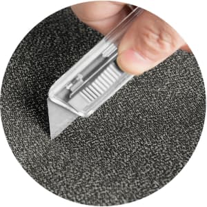 A hand holding a utility knife against a textured grey surface. The knife features a retractable blade.