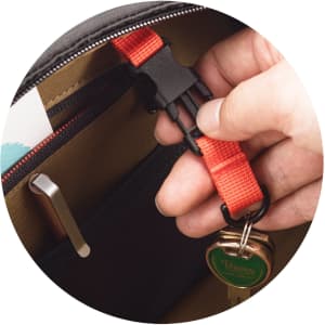 A hand holding a red strap clip inside a black and brown bag, with a keychain and various items visible.