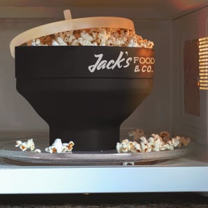 A black microwave popcorn popper with popcorn overflowing, located inside a microwave. It features a logo.