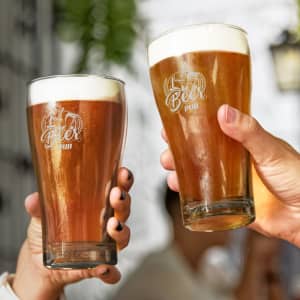 Two clear glass schooner beer glasses filled with amber beer and topped with creamy white foam, featuring a logo.