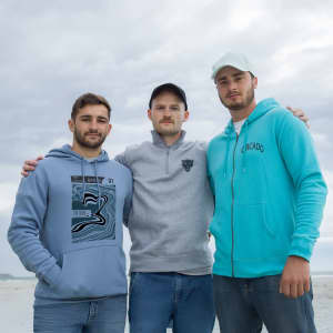 Three men stand together wearing hoodies in various shades of blue and grey. One hoodie features a logo.