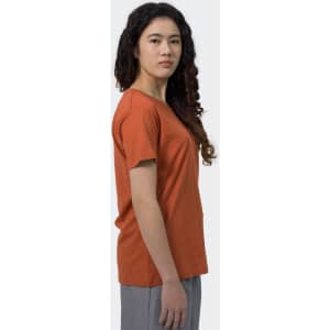A woman in a rust-coloured T-shirt stands sideways, with curly hair and a neutral expression. The shirt features a logo.