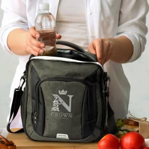 A grey cooler bag being filled with a drink bottle, accompanied by fresh food items on a wooden surface.