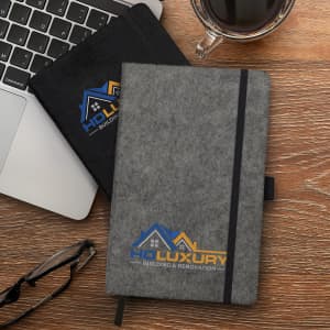 A grey RPET felt hard cover notebook with an elastic band, next to a black version and a coffee cup on a wooden surface.