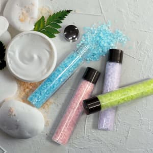 A set of bath salts in clear tubes featuring blue, pink, green, and lavender colours, alongside a white cream in a jar and pebbles.
