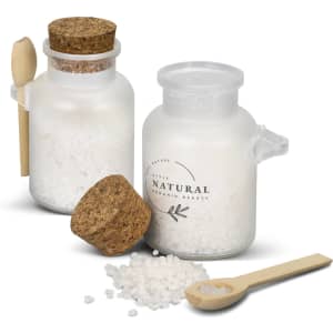 A glass jar containing white bath salts with a cork lid and a wooden spoon nearby. The jar has a simple branding design.