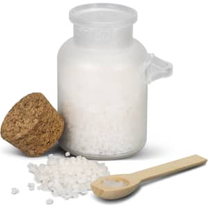 A clear glass jar filled with white bath salts, topped with a cork lid and accompanied by a wooden spoon.