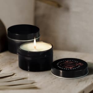 A black travel candle with a lit wick, placed beside its matching lid on a neutral surface. It features a logo on the lid.