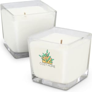 A square, white scented candle in a clear glass container, featuring a logo design. Two candles are shown.