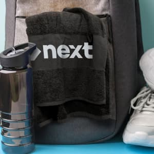 A black gym towel with a logo hangs from a grey backpack, next to a drink bottle and white running shoes.