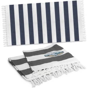 A striped cotton towel in navy blue and white, with a smaller grey and white striped towel underneath. Both towels have tassels and feature a logo.