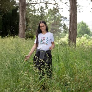 A woman in a white T-shirt and black pants stands in tall green grass, surrounded by trees. The T-shirt features a logo.