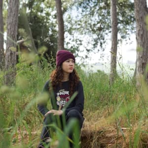 A woman with curly hair sits on a log in a forest, wearing a black long sleeve crew shirt and a burgundy beanie.