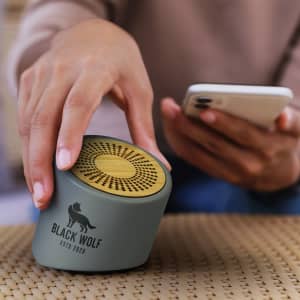 A green and gold NATURA Limestone Bluetooth mini speaker is being held next to a smartphone on a textured surface.