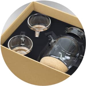 A tea set featuring a clear glass teapot with cork base and two matching glass cups, presented in a brown cardboard box.