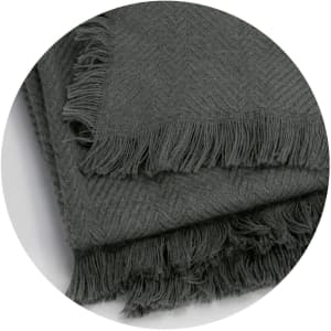 A grey throw blanket featuring a herringbone pattern and fringed edges, displayed in a circular format.