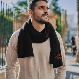 A man wearing a black brushed scarf, standing outdoors with palm trees in the background. The scarf features a logo.