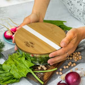 A glass food container with a bamboo lid, surrounded by fresh vegetables like spinach and radishes.