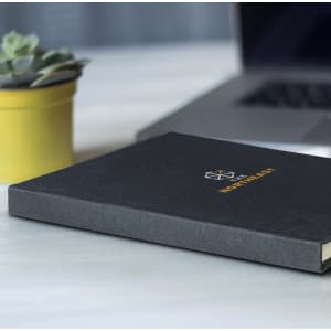A recycled cotton hard cover notebook in black, placed on a desk next to a yellow plant pot and a laptop.