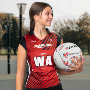 A woman in a red and black netball bib holds a netball, with a sports court in the background.