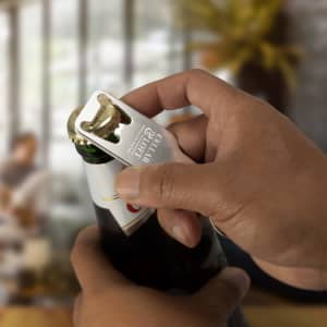 A drink bottle opener key ring in silver being used to open a green bottle. The opener includes a logo.
