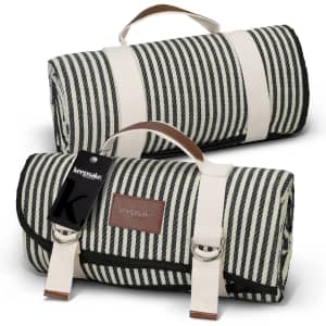 A striped picnic blanket in black and cream, folded with handles and a tag. It features a brown leather label.