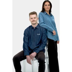 A denim shirt in two shades of blue, worn by a man sitting and a woman standing. The shirts feature a logo.