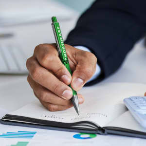 A metal pen in green is being held above an open notebook, with a hand writing notes. The scene includes a calculator and graphs.