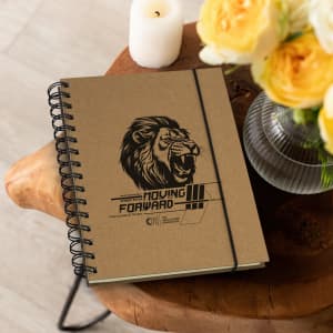A brown spiral notebook with a lion graphic and a logo, placed on a wooden table beside yellow flowers and a candle.