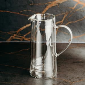 A drink jug made of clear glass with a curved handle and spout, featuring a sleek, modern design.