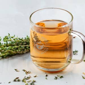 A glass mug with a transparent design, containing a herbal drink, beside fresh thyme sprigs on a light surface.
