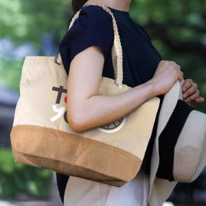 A tote bag in cream with a textured brown base, featuring a logo and being held by a person wearing a hat.