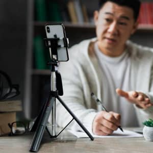 A black selfie stick tripod holds a smartphone, with a person writing in the background. The setting is casual.