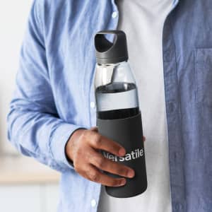 A drink bottle with a transparent glass body and a black silicone sleeve, held by a person wearing a blue shirt.