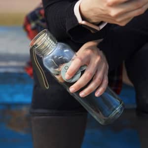 A clear drink bottle with a metal lid and a green silicone strap, held by a person sitting down.