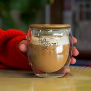 A glass cup with a wooden lid, featuring a frothy coffee inside, held in a hand with a red sweater.