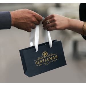 A small black paper bag with a white ribbon handle, featuring a logo in gold.