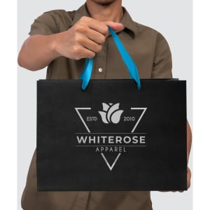 A person holds a large black paper bag with a blue ribbon handle and a logo on the front.