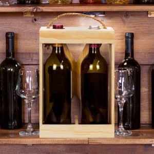 A wooden wine crate in natural finish holds two wine bottles with a rope handle, surrounded by glasses and other bottles.