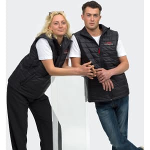 A black unisex puffer vest is worn by two models, one leaning against a clear display stand, showcasing its design.
