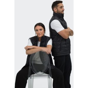 A black puffer vest featured on two models, one seated on a transparent chair and the other standing behind.