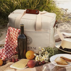 A beige cooler tote bag with a logo, a drink bottle, cheese, fruit, crackers, and a checkered cloth on grass.