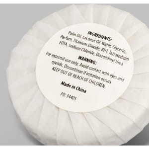 A white circular soap wrapped in paper, labelled with ingredients and warnings, features a logo.