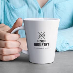 A white coffee mug with a logo, held in a hand, on a grey surface, with a blue shirt in the background.